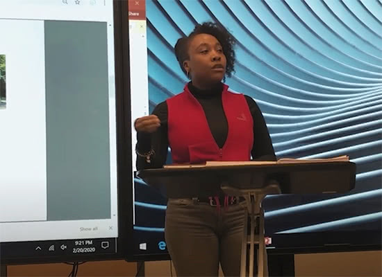 A female student speaks at a podium with a presentation projected behind her