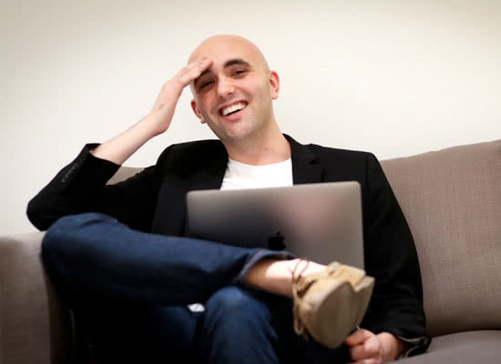 T.J. Kelly smiles while sitting on a couch with a laptop on his lap