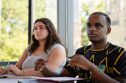 Students listening to a lecture in a classroom with large glass windows and lots of natural light