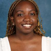 Sarah Naje smiles and poses in front of a blue background wearing a white blouse 