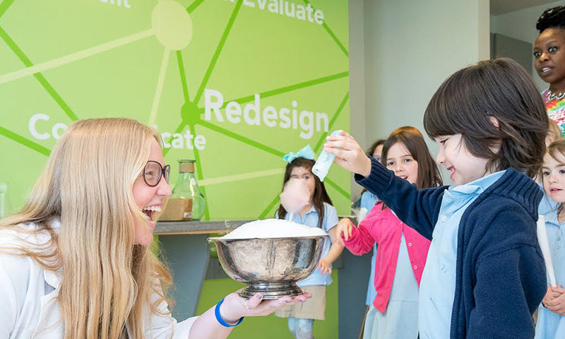 A kid doing a science experiment with a woman while other kids in the background watch