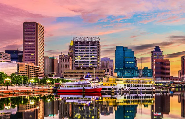 Baltimore skyline with skyscrapers, the harbor, and a boat with blue and pink skies at dusk
