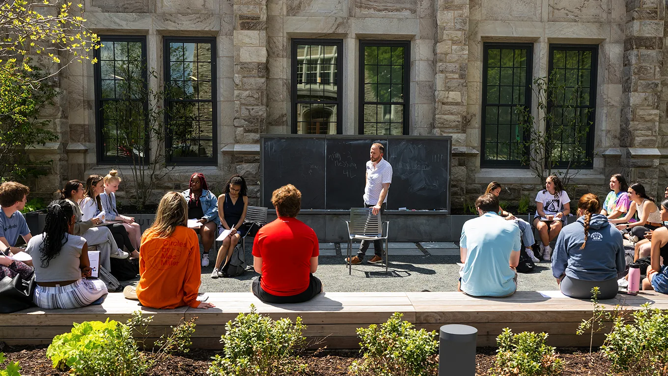 Students and a professor sit on the grass in a circle while having an outdoor class - Press enter to play