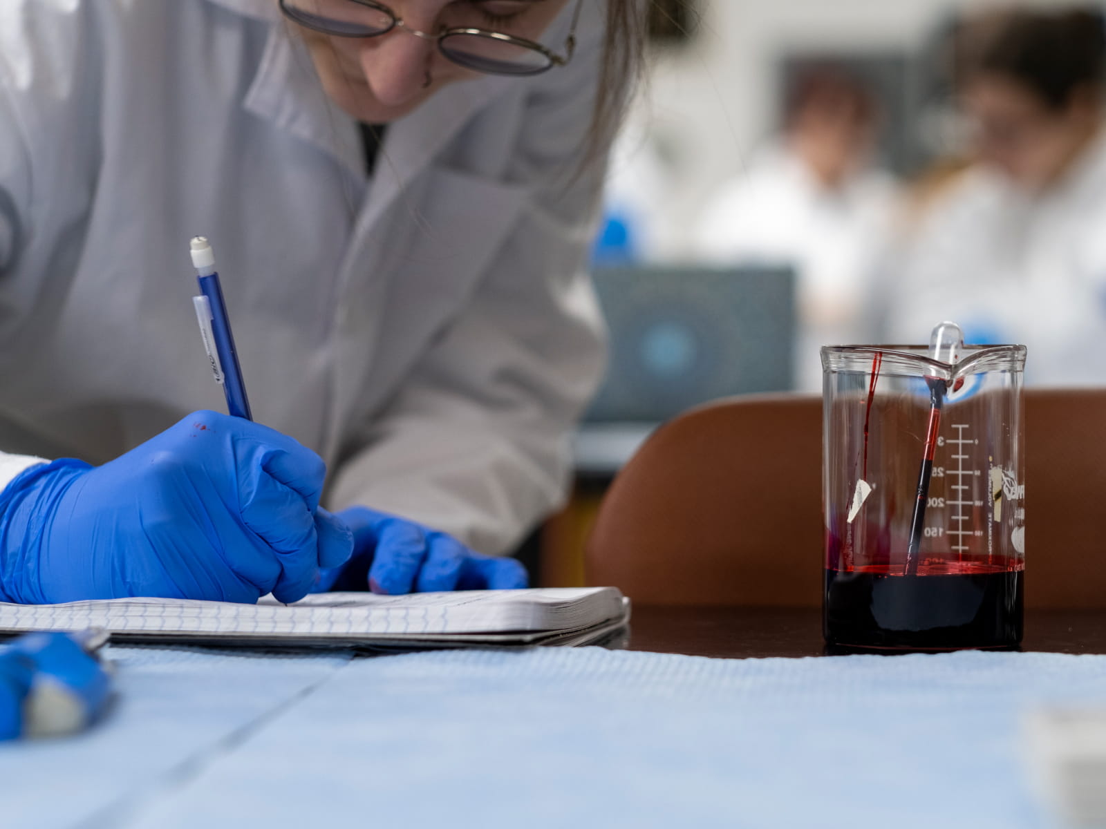 A student in a white lab coat and gloves writes on a counter next to a beaker of blood