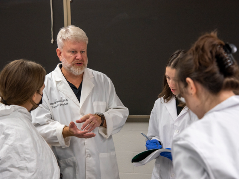An instructor in a white lab coat speaks to a small group of students taking notes