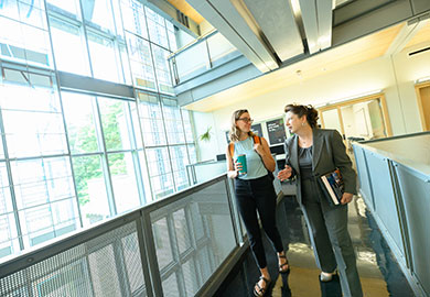 Student walking the footbridge inside a building as the sun shines through the vast glass facade