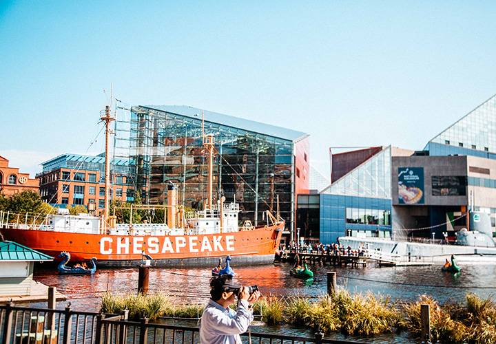 A view of the Inner Harbor on a sunny day, featuring the aquarium, a large sailboat, dragon pedal boats, and a tourist taking photos