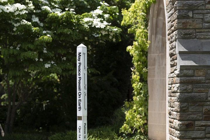 A white pole with the words "May Peace Prevail on Earth" stands by a stone wall