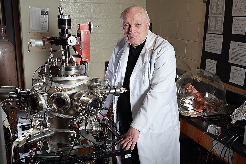 Fr. Frank Haig, S.J., stands in his physics lab wearing a lab coat and smiling