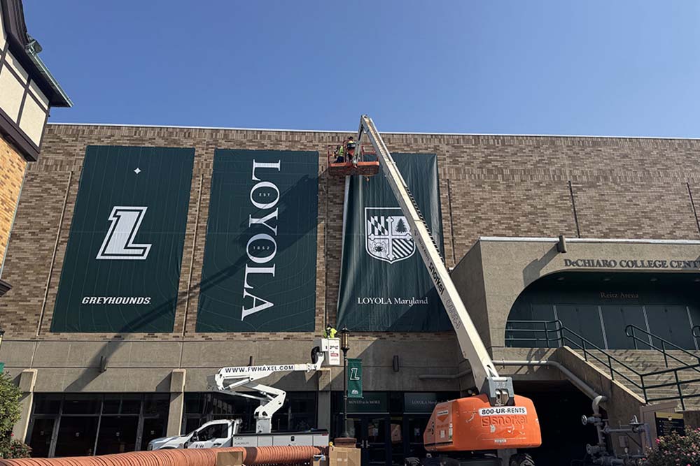 Workers on lifts hanging Loyola Maryland and Greyhounds banners on the Loyola DeChiaro College Center