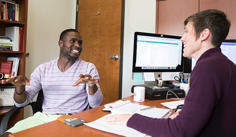 A student sitting across the desk from a professor talking and smiling