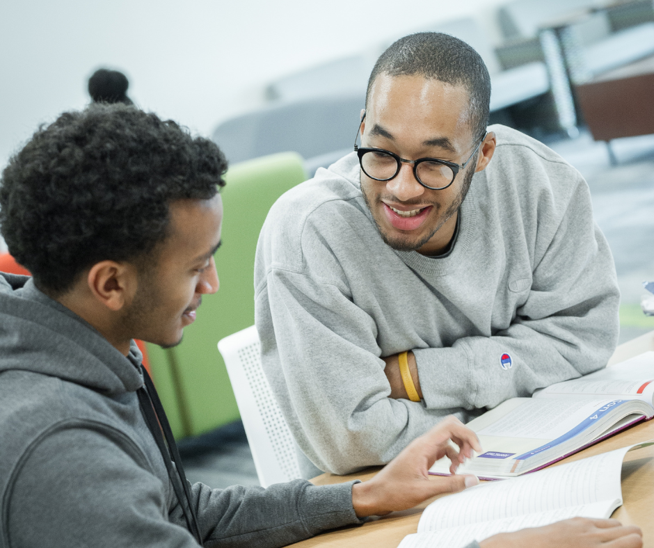 Two students work together on homework, at a table with papers.