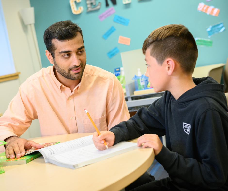 A male teacher works at a table with a male student on work.