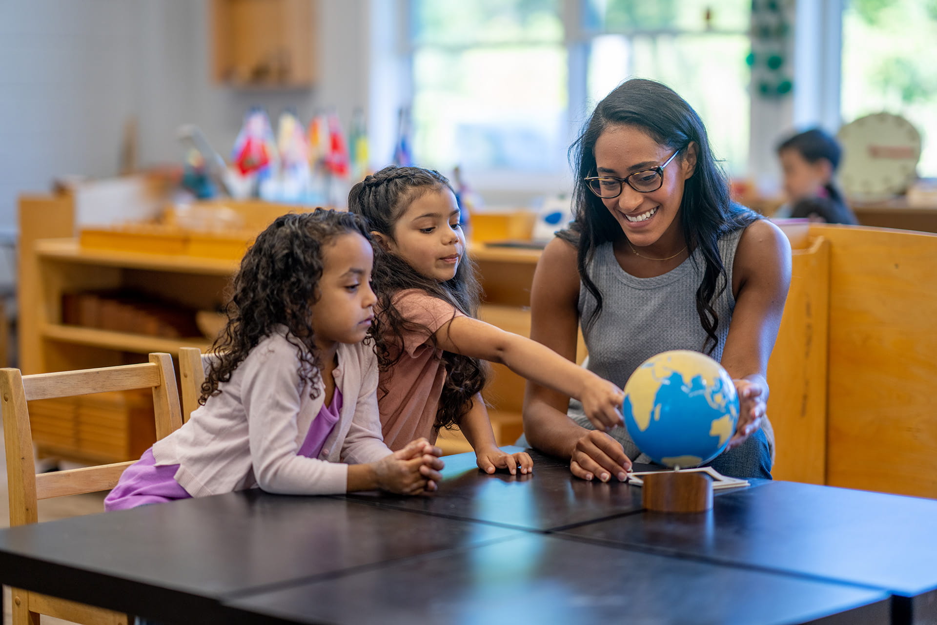 A female teacher sitting at a desk with students looking at a globe.