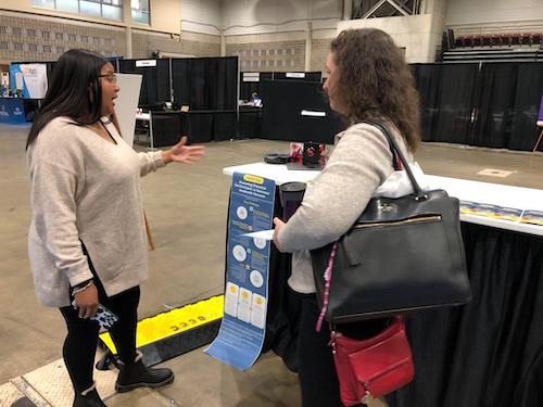 Two woman standing in fornt of a table with a research poster, one of the women explaining the poster to the other