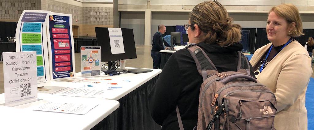 Two women, one in a blazer and the other wearing a backpack looking at a poster presentation on a table