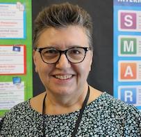 Joanna Edwards, woman with short dark hair and glasses in front of educational posters in a classroom