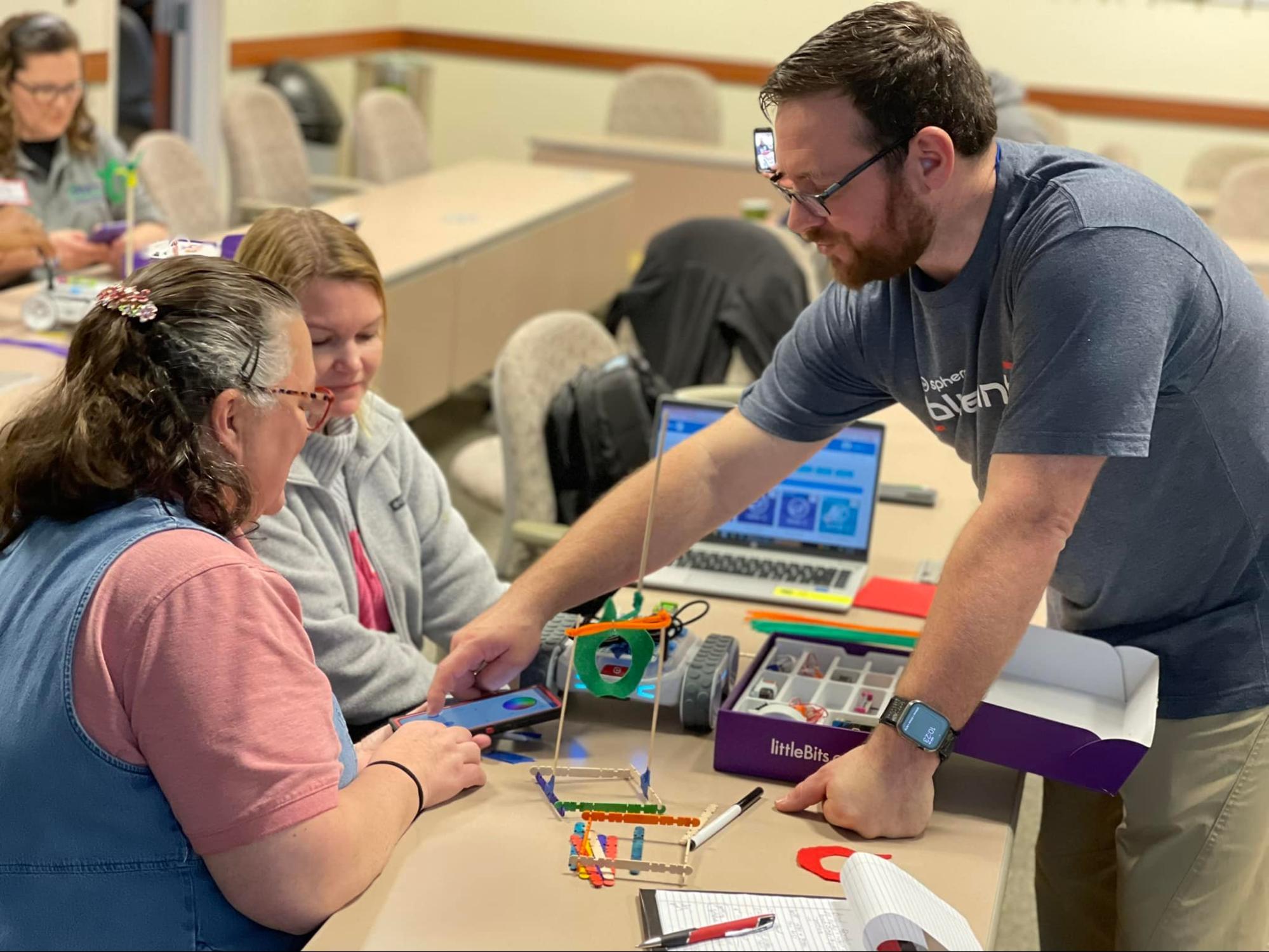 Two women seated and one man standing, collaborating to code their RVR+ robot and build the littleBits components