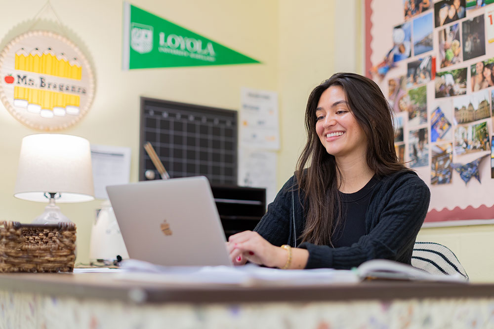 Teacher smiles while sitting at desk working on laptop.