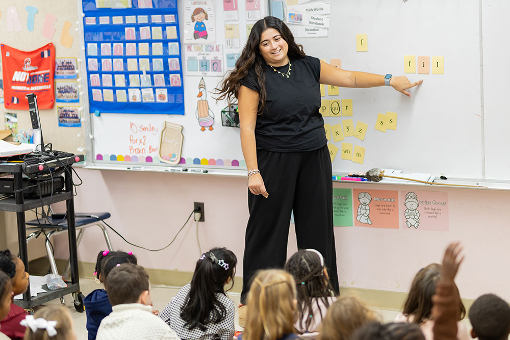 Teacher standing at front of class pointing to letters on the board.