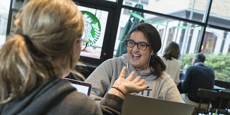 Two female students chatting while on laptops at the campus Starbucks