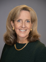 Trish Tarrant smiles for a portrait with a gold necklace and earrings in front of a gray backdrop