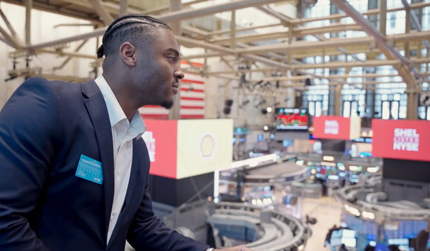 A young man in a dark colored suit looks over the balcony at the New Your Stock Exchange.
