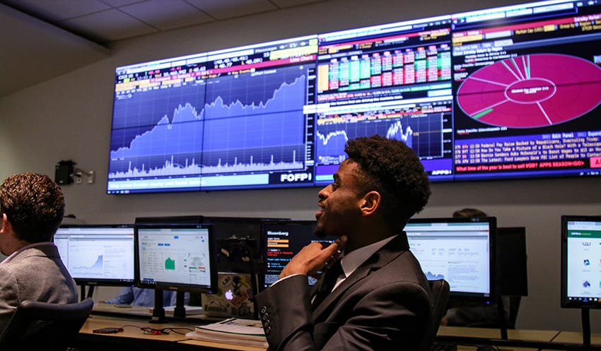 A student in a suit sits in front of a computer and a video wall with data visualizations including a pie chart and bar graph.