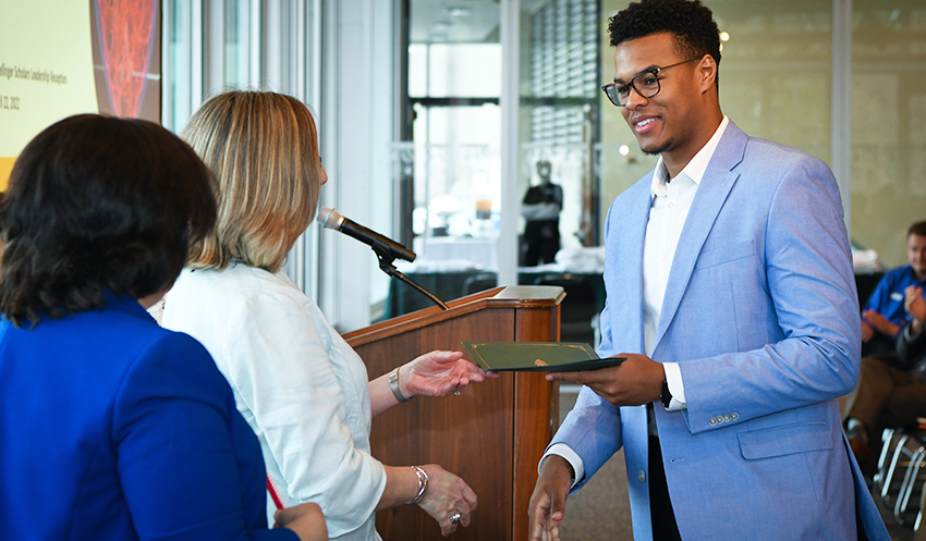 A student wearing glasses and a suit receives documents from two professors