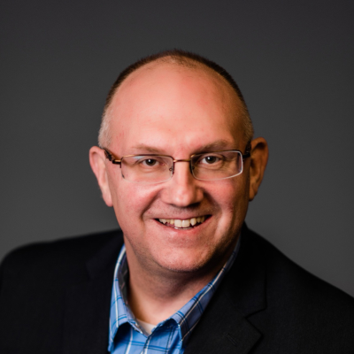 Professional headshot of Bill Harmon, depicting a smiling middle-aged man wearing glasses, a blue checkered shirt, and a dark blazer against a neutral studio background.