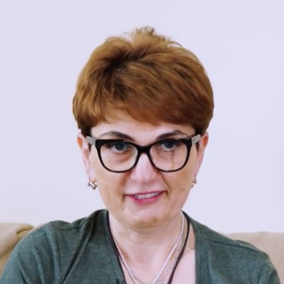 Photo of Russo Tsereteli, depicting a smiling middle-aged woman wearing glasses, a green Loyola University shirt, sitting on a couch with a laptop