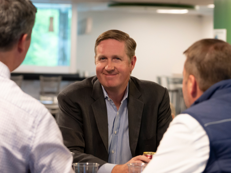 Photo depicting a middle-aged man smiling and looking into the cameras, wearing a blue striped shirt, and a dark blazer in a social setting. Flanked by a man on each side, both with their backs to the camera. The man on the right is wearing a white shirt and a blue vest, the one on the left is wearing a light checkered button down shirt