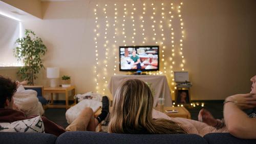Roommates watch TV in a dorm room decorated with string lights and a plant