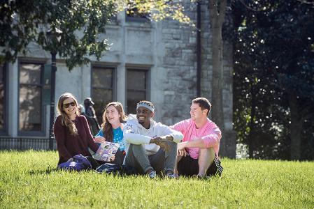 A group of students sit on the grass on the quad and laugh as they enjoy the sunshine.