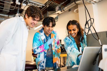 Three students smiling and discussing lab work using a laptop.