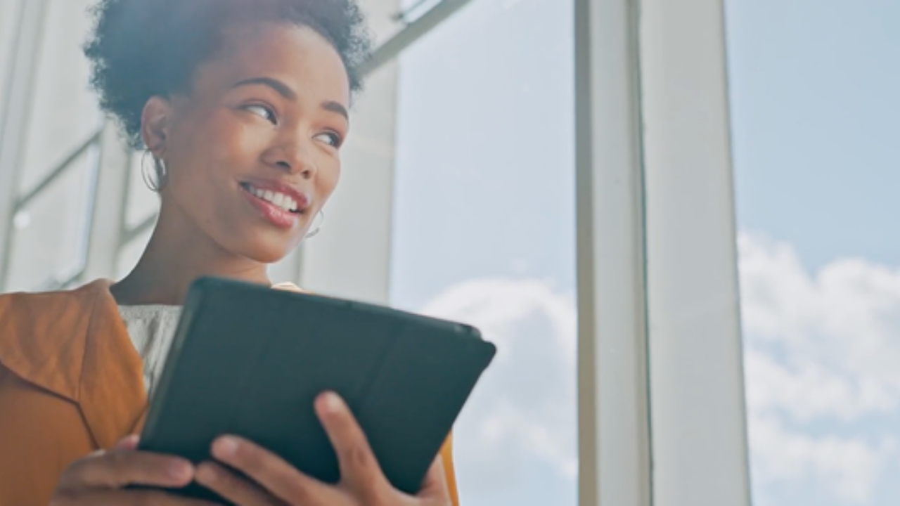 A woman holding a tablet smiles while gazing out a large window. - Press enter to play