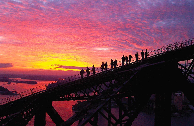Silhouette of students on a bridge in front of a bright pink, purple, and yellow sunset