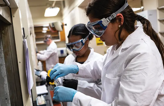 Students wearing white lab coats and eye protection while working with chemicals