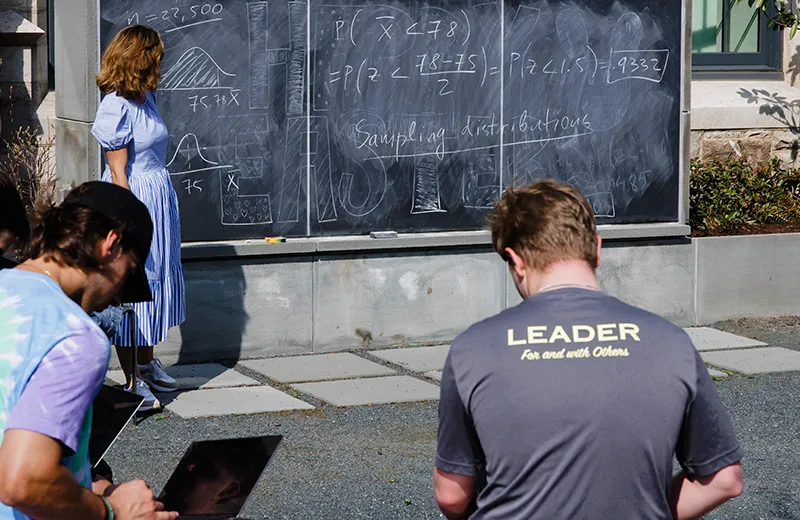 A student with a shirt that reads "Leader for and with others" sits in front of an outdoor chalkboard