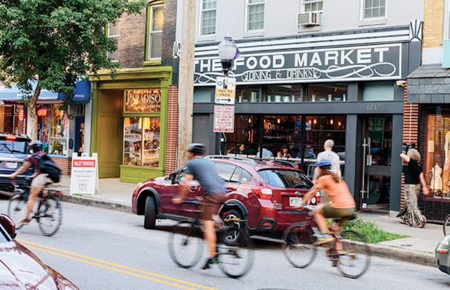 Bicyclists ride down a street in Hampden in front of The Food Market
